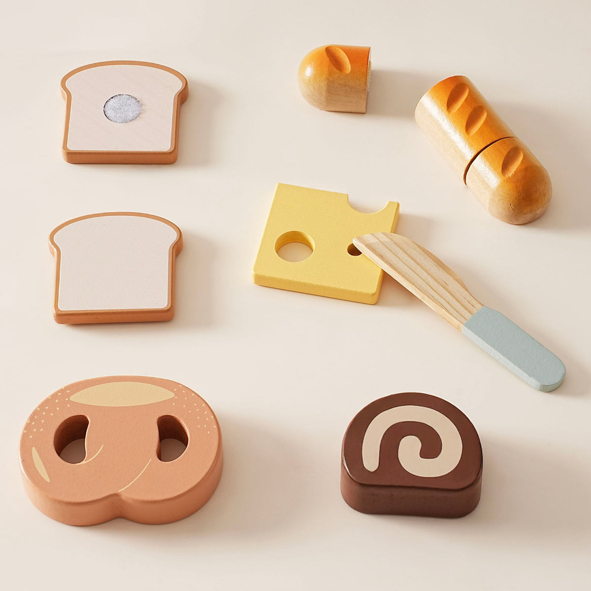 ROBUD wooden bread and dessert cutting playset pieces flat lay on cream background showing toast halves, yellow cheese, baguette halves, pretzel, swiss roll, and wooden knife