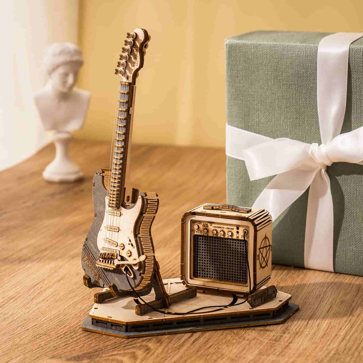 ROKR electric guitar 3D wooden puzzle with mini amp displayed beside a ribbon-wrapped gift and marble bust
