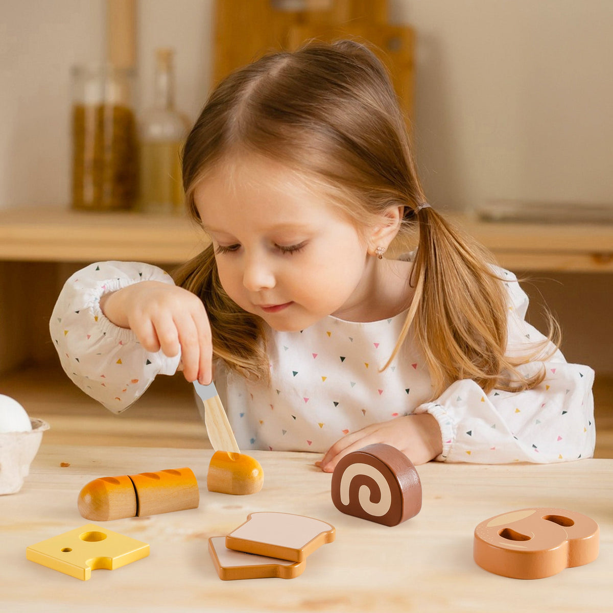 Young girl cutting a wooden baguette with the ROBUD bread cutting playset on a wooden table with swiss roll, toast, cheese, and pretzel nearby