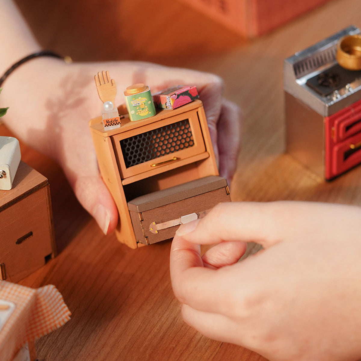 Rolife Cozy Kitchen miniature hands assembling a tiny wooden oven unit with colorful canned goods on top