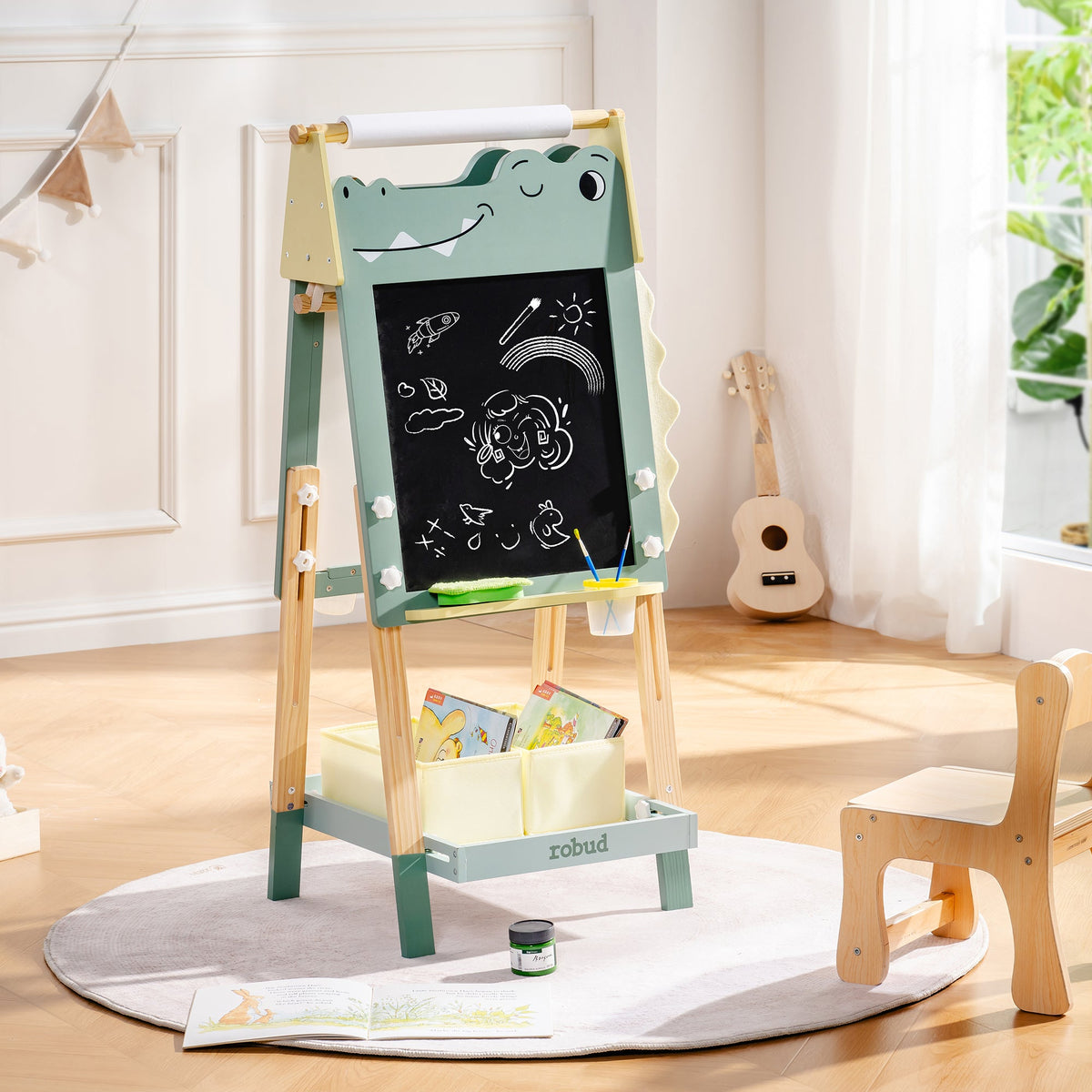 ROBUD green dinosaur double-sided chalkboard easel with storage bins and paper roll shown in a cosy play corner.