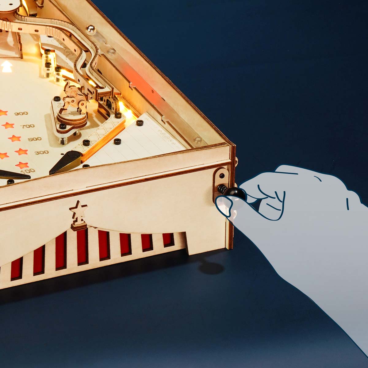Side view of the ROKR 3D Pinball Machine wooden puzzle showing a hand pulling the spring-loaded plunger knob to launch the metal ball into the lit playing field