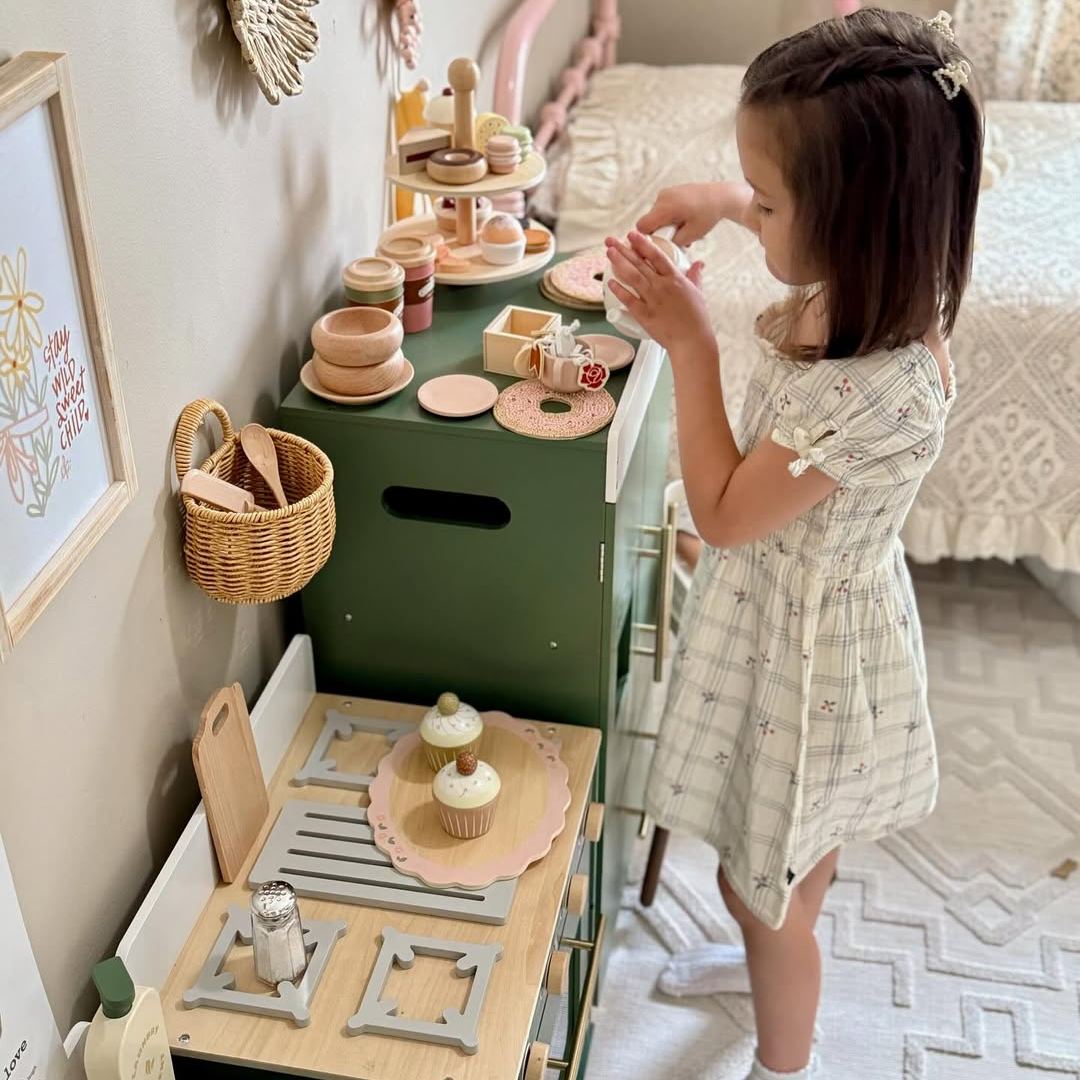 Young girl in floral dress playing with the ROBUD green play oven and kitchen set in a cozy room with wicker basket accessories