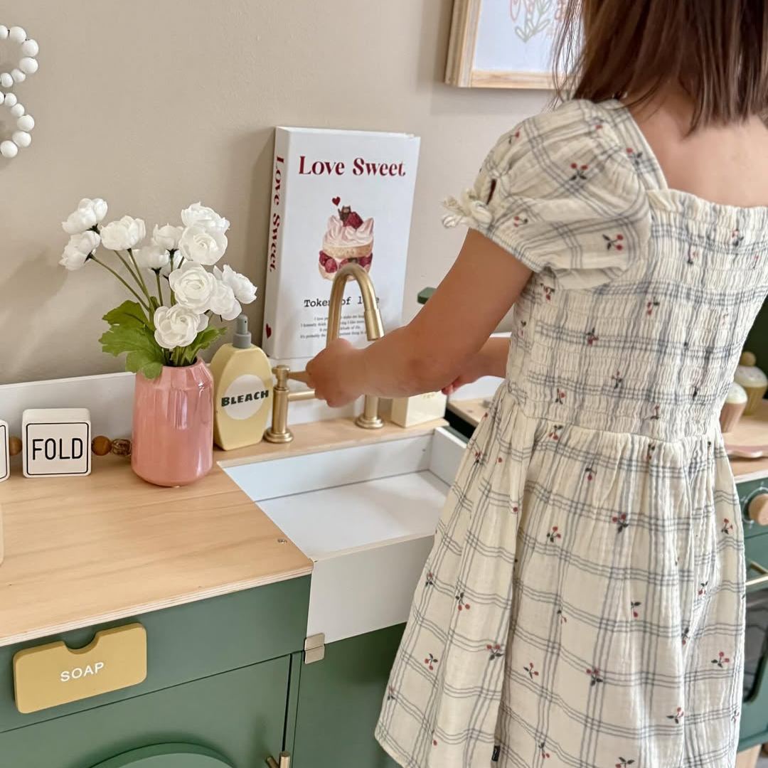Girl in floral dress reaching toward the ROBUD vintage green play kitchen sink area with cookbook and flower arrangement nearby