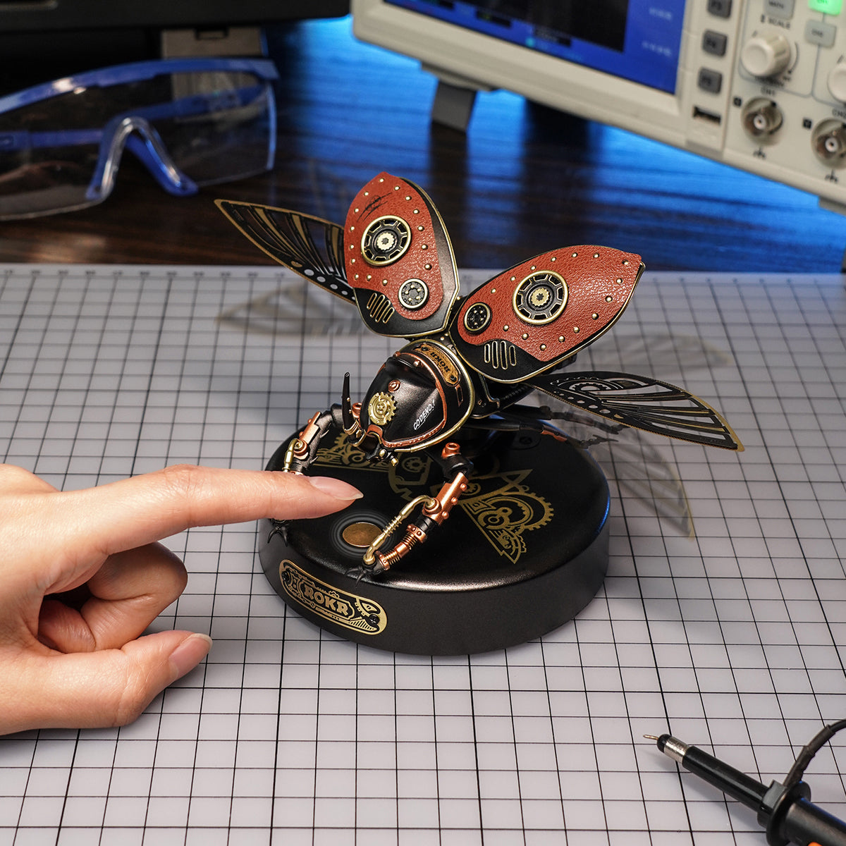 ROKR steampunk metal beetle model on electronics workbench with oscilloscope and finger pointing at it